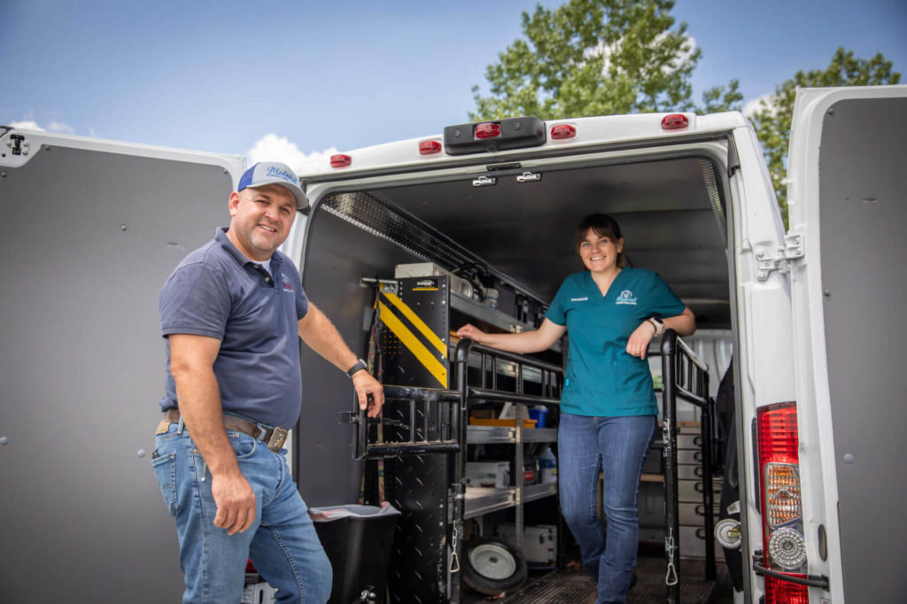 A man and woman smile at the camera while loading equipment into the back of a service van.